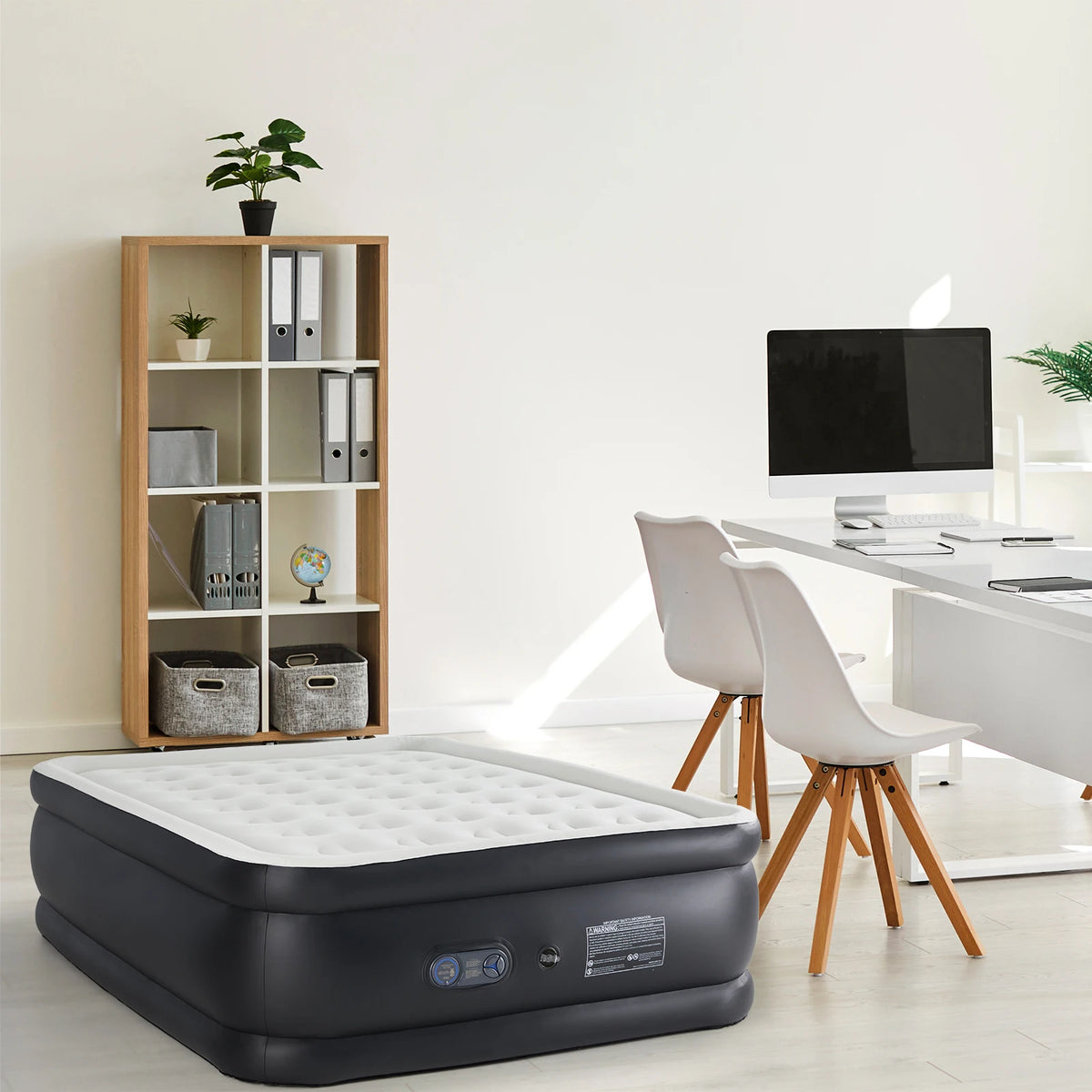A black elevated air mattress with a white top is placed on a light floor next to a wooden shelf and a white desk.