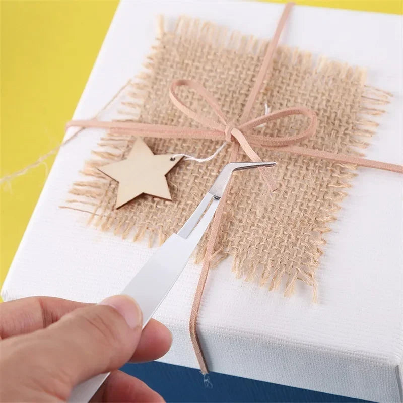 A hand holds a silver vinyl weeding and craft tool set with a star charm tied to burlap on a white gift box.