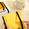 A yellow sketchbook is secured with a black elastic strap, shown next to a white mug.