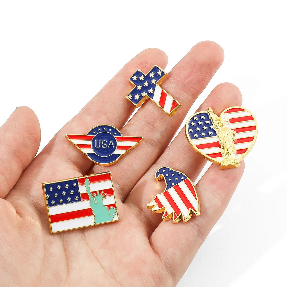 A hand holds several enamel pins featuring the American flag design, including a cross, an eagle, and stripes.