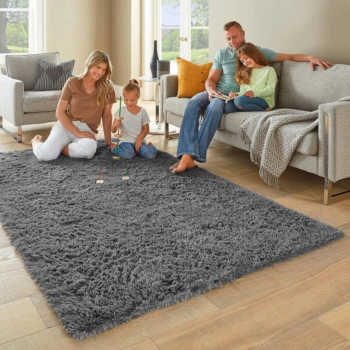 Eine Familie sitzt auf einem grauen flauschigen Teppich in einem Wohnzimmer mit Sofa und großen Fenstern.