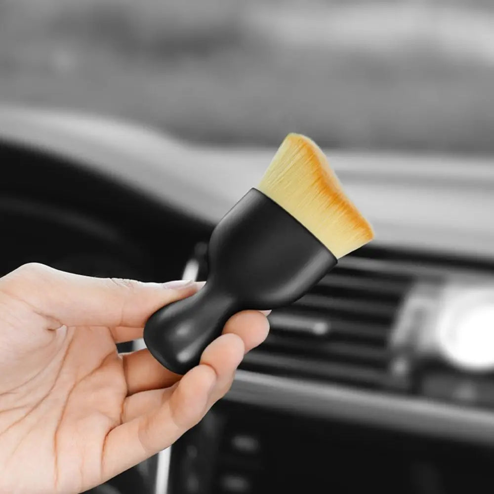 A hand holds a black detailing brush with a yellow curved bristles, positioned near a car vent.