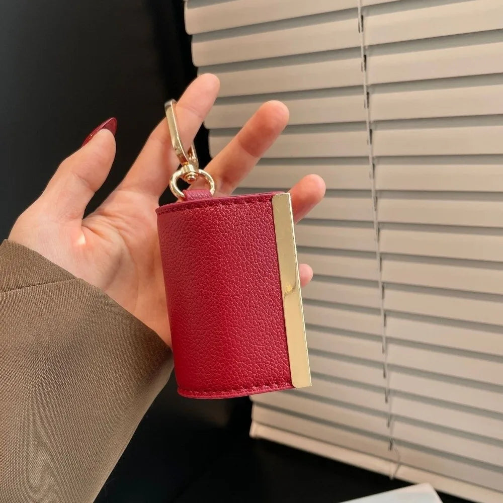 A hand holds a red leather keychain tote bag with a gold clasp against a light gray slatted blind.
