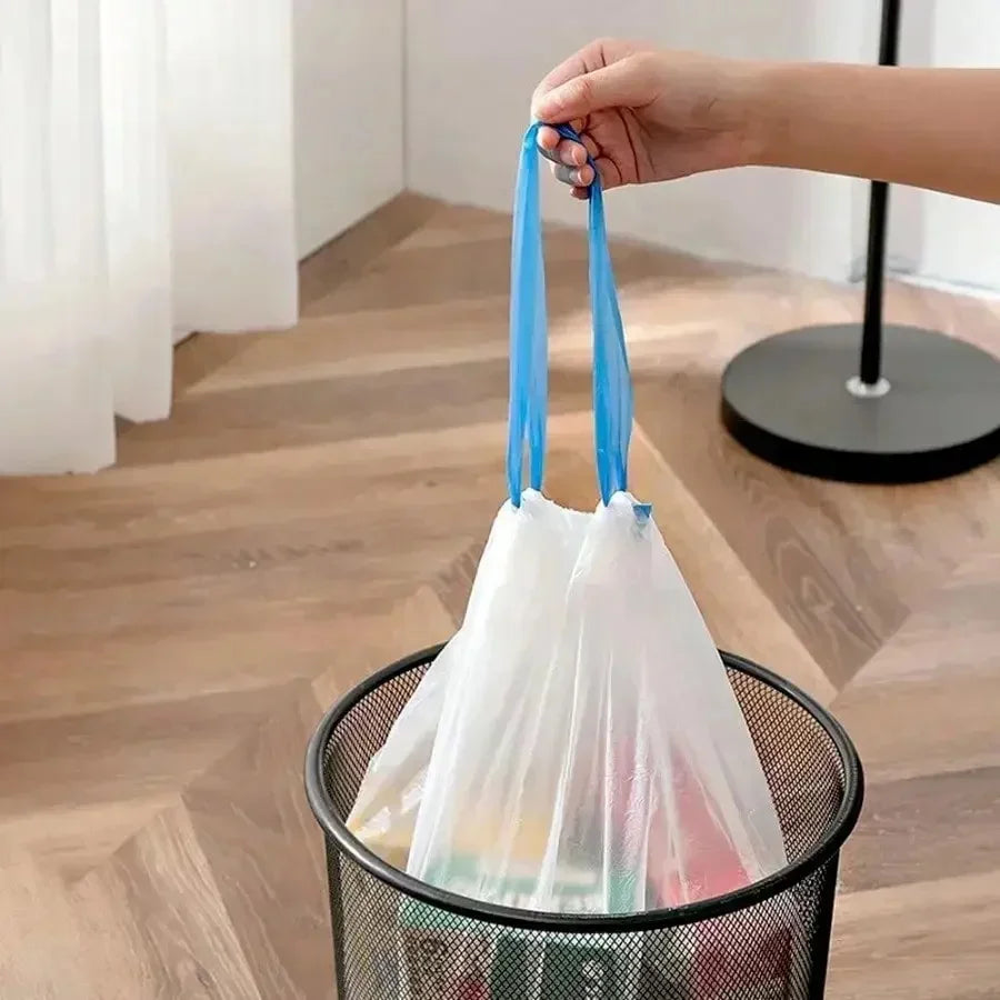 A hand is holding a white drawstring trash bag with blue handles over a black mesh basket.
