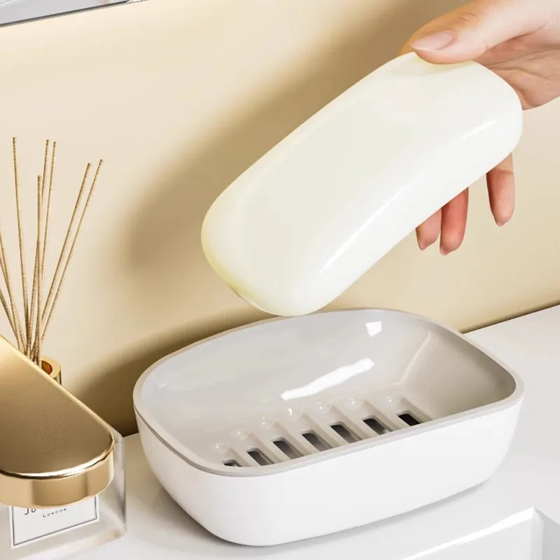 A hand is holding a white double-layer draining soap dish with a non-slip base against a light background.