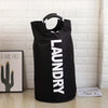 A black foldable laundry bag with a padded handle is displayed against a white brick wall.