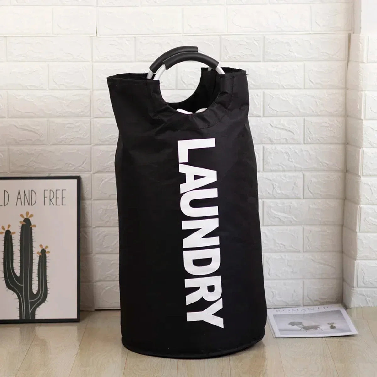 A black foldable laundry bag with a padded handle is displayed against a white brick wall.