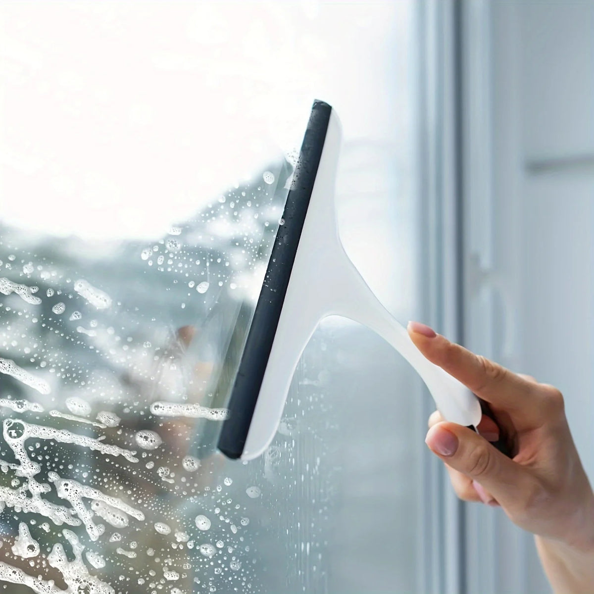 A hand uses a squeegee to clean water droplets off a window pane.
