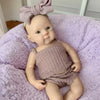 A handcrafted silicone reborn doll with a pink bow is lying in a purple fluffy blanket.
