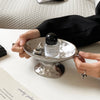 A hand holds a silver ceramic soap dish with an elevated stand and a glass bottle of perfume.