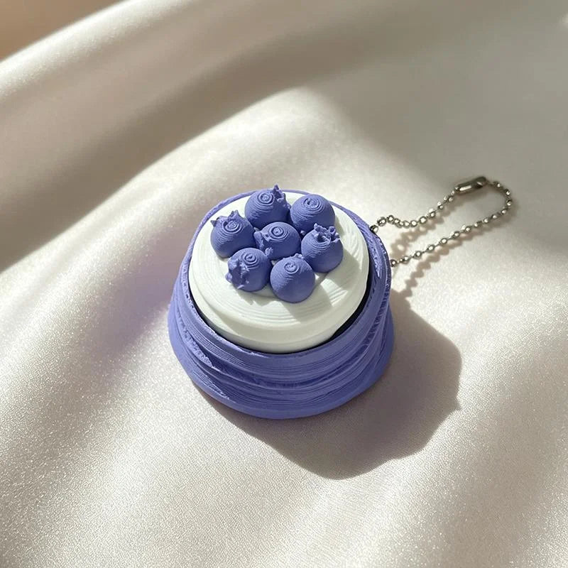 A small purple and white layered cake with blue fondant flowers is presented as a fidget keychain.