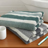 A stack of green and white striped quick-dry bath towels next to a white cup and a blue toothbrush on a wooden surface.