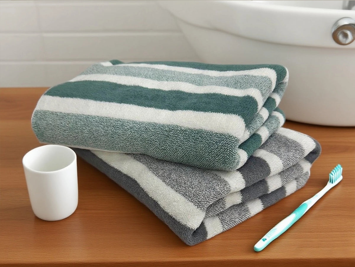 A stack of green and white striped quick-dry bath towels next to a white cup and a blue toothbrush on a wooden surface.