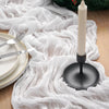 A white crinkle gauze table runner draped over a wooden surface with a tall white candle and festive greenery.