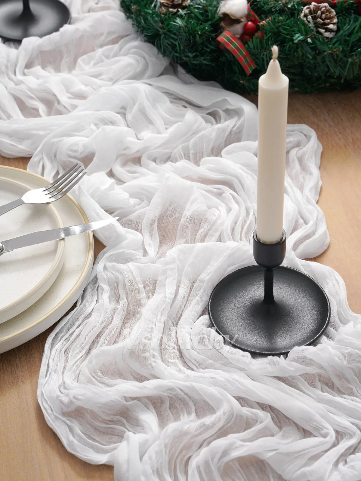 A white crinkle gauze table runner draped over a wooden surface with a tall white candle and festive greenery.