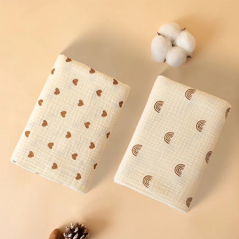 A soft cotton gauze baby swaddle blanket with a subtle pattern is shown next to a fluffy white pom-pom.