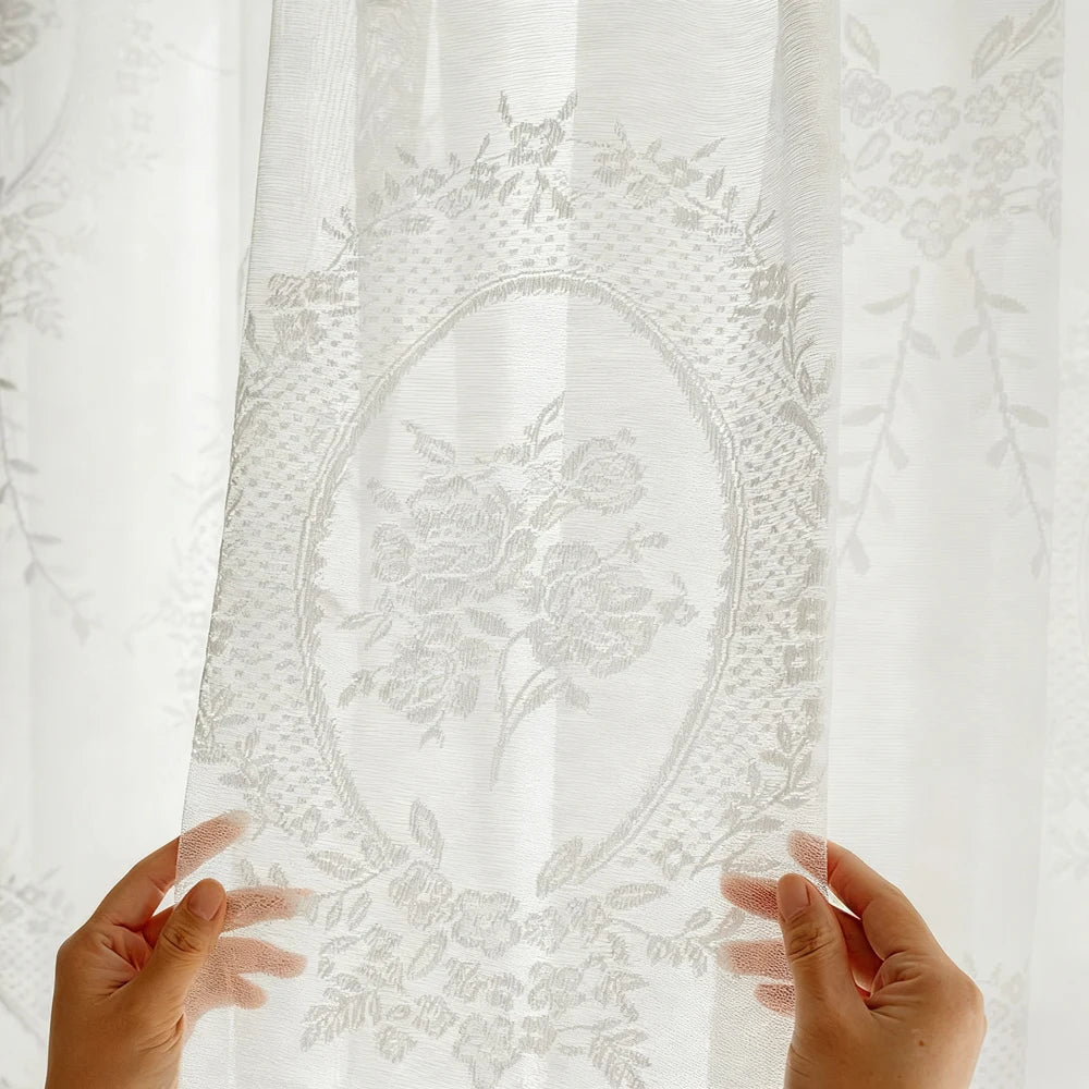 Hands holding a sheer white lace curtain with floral embroidery, showing its delicate texture and light filtering quality.