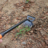 A black and orange 2-in-1 stainless steel weeding shovel and rake is shown in dirt and leaf litter.