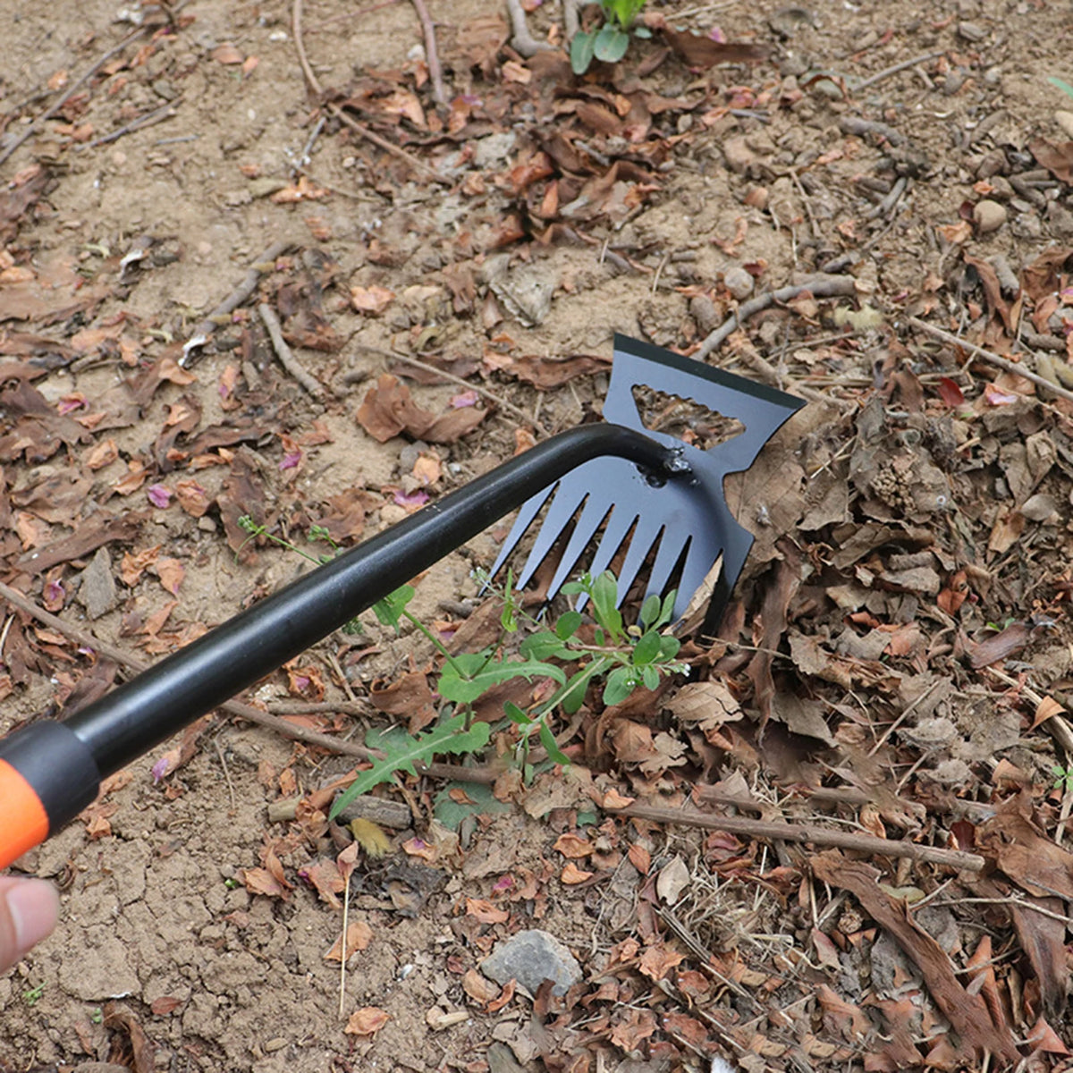 A black and orange 2-in-1 stainless steel weeding shovel and rake is shown in dirt and leaf litter.