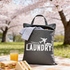 A black laundry bag with a white graphic is placed on a checkered blanket in an outdoor spring setting.