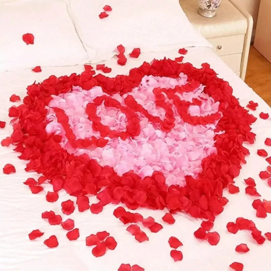 A heart shape made of red and pink silk rose petals scattered on a white bed.