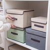 A hand is placing a beige fabric storage box onto a green and gray storage box set inside a white shelf.