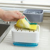A hand is placing a yellow sponge into a white kitchen soap dispenser with a blue base and a gray storage basket.