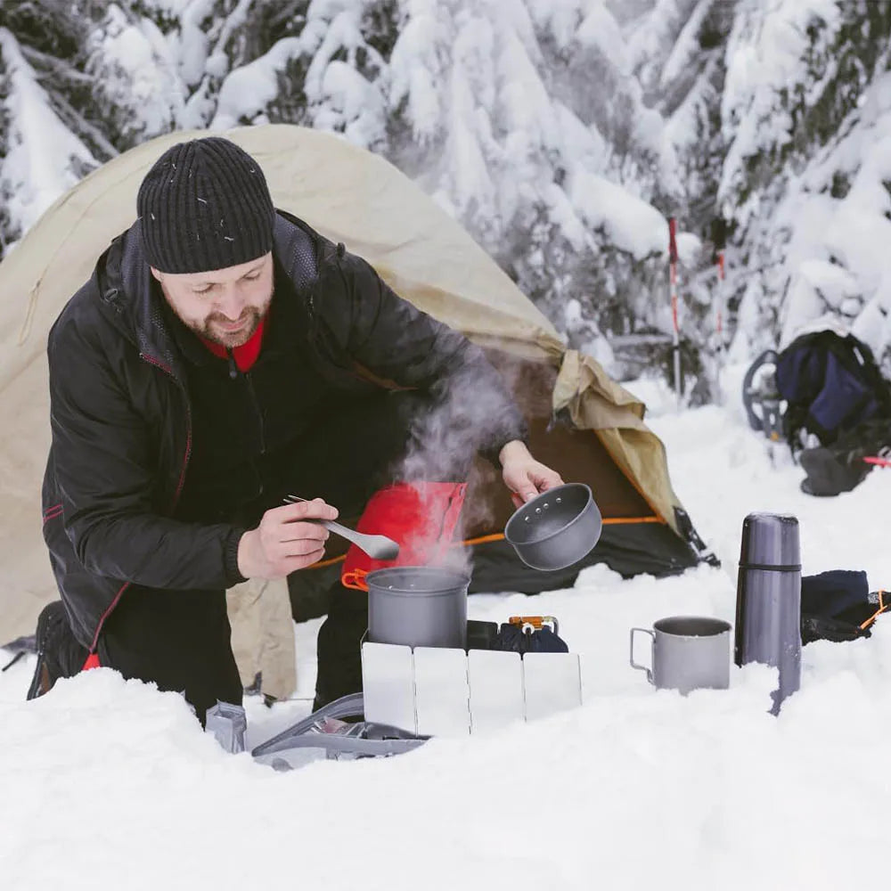 A person is cooking in a pot outdoors next to a tent in the snow using an adjustable aluminum camping stove.