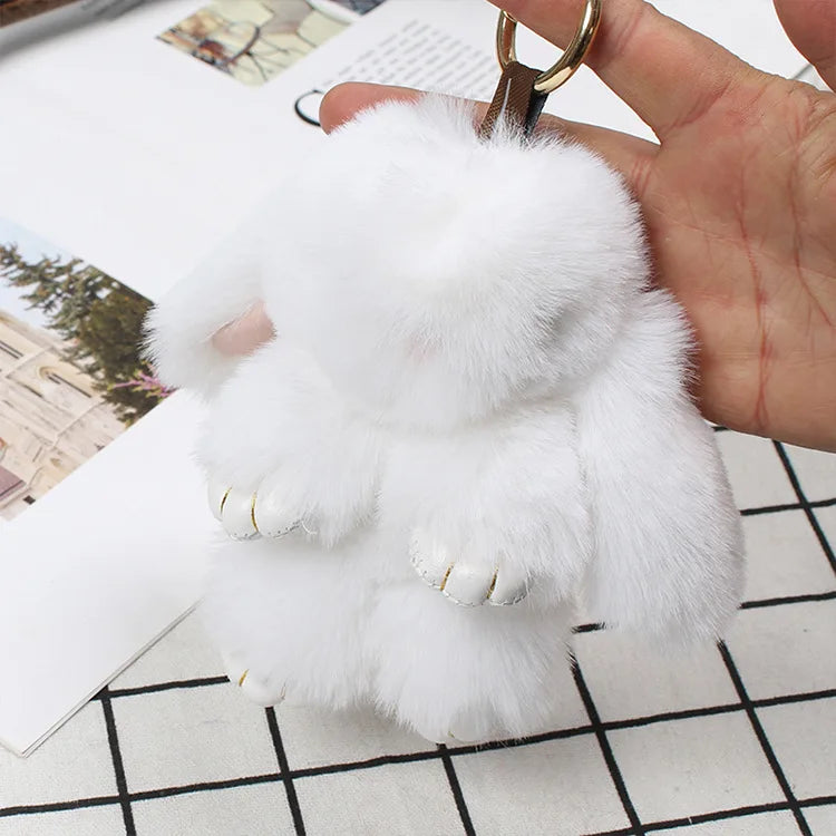 A white fluffy angel bunny keychain with a pom pom tail is held by a hand against a light background.