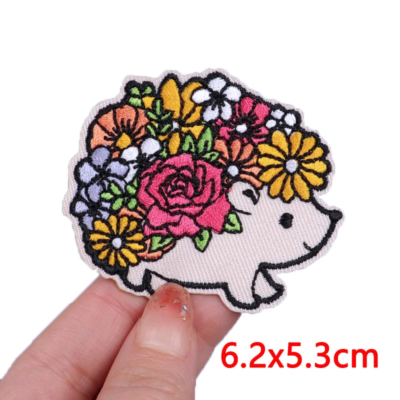 A hand holds a colorful embroidery patch featuring a pink and yellow floral design surrounding a cute white cartoon bear face