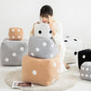 A person is holding a white polka-dotted plush pillow next to several gray and tan colored soft cushions.