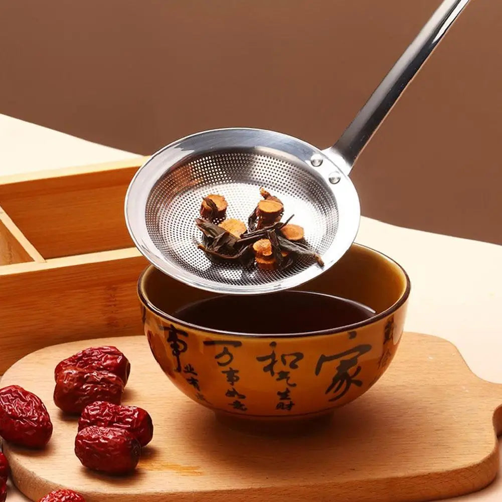 A stainless steel fine mesh skimmer spoon is shown above a small bowl of dark liquid and several dried red fruits on a wooden