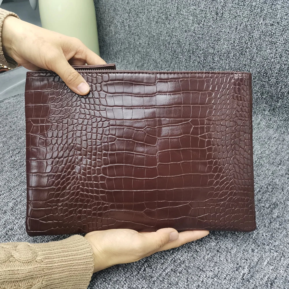 A hand holds a dark brown crocodile-embossed PU leather envelope clutch against a gray fabric background.
