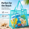 A turquoise breathable mesh tote bag filled with beach items like towels, toys, sandals, and sunscreen on a sandy beach.