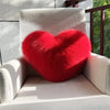 A bright red, fluffy heart-shaped throw pillow resting on a light-colored armchair.