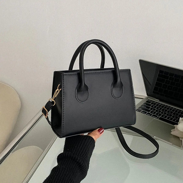Bolso cruz negro clásico de cuero PU con herrajes dorados sobre una superficie de vidrio.
