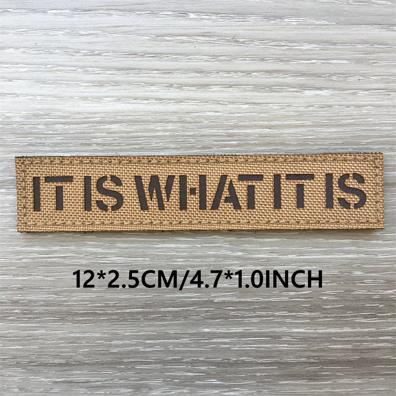 A brown woven patch with the text IT IS WHAT IT IS engraved on it, measuring 12x2.5cm or 4.7x1.0inch.