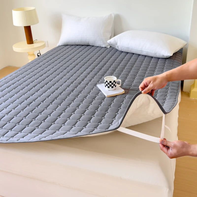 Grauer gesteppter wasserdichter Matratzen-Schutz auf einem Bett mit Kissen und einer Tasse.