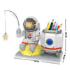 A LEGO astronaut figure is shown with a light beam, standing on a yellow base, next to colored pencils.