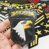 A hand holds a black embroidered patch featuring a white eagle head on a dark background, surrounded by other military-themed
