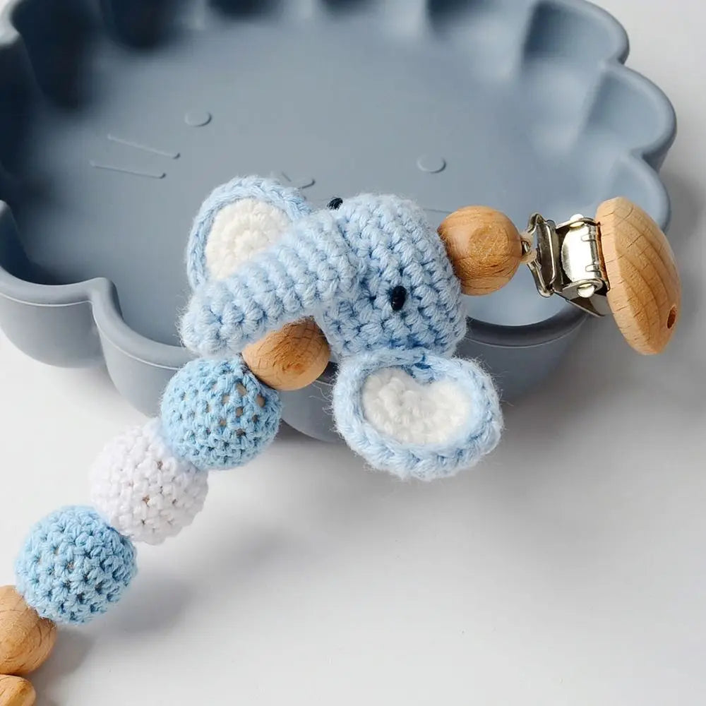 A wooden pacifier clip made of light blue crochet beads with a wooden attachment, resting in a gray dish.
