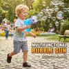 A young boy is playing with a blue bubble gun, shooting bubbles into the air in a park setting.
