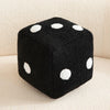 A black dice-shaped plush pillow with white polka dots resting on a light surface.