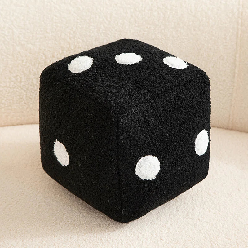 A black dice-shaped plush pillow with white polka dots resting on a light surface.