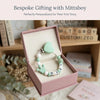 A pink box containing a personalized baby pacifier clip with a green heart design and custom name