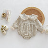 A beige floral lace cotton romper for a baby girl is displayed next to wooden mats and white accessories.