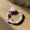 A pink beaded bracelet with a heart charm and custom name beads lying on a wooden surface.
