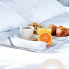 A white linen surface features a cup of coffee, an orange juice glass, and a stack of croissants with a folded striped kitche