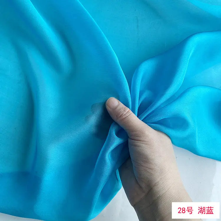 A hand is folding a piece of bright cyan blue silk chiffon fabric.