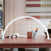 A white half-moon LED arc desk lamp illuminates a workspace with art supplies and books.